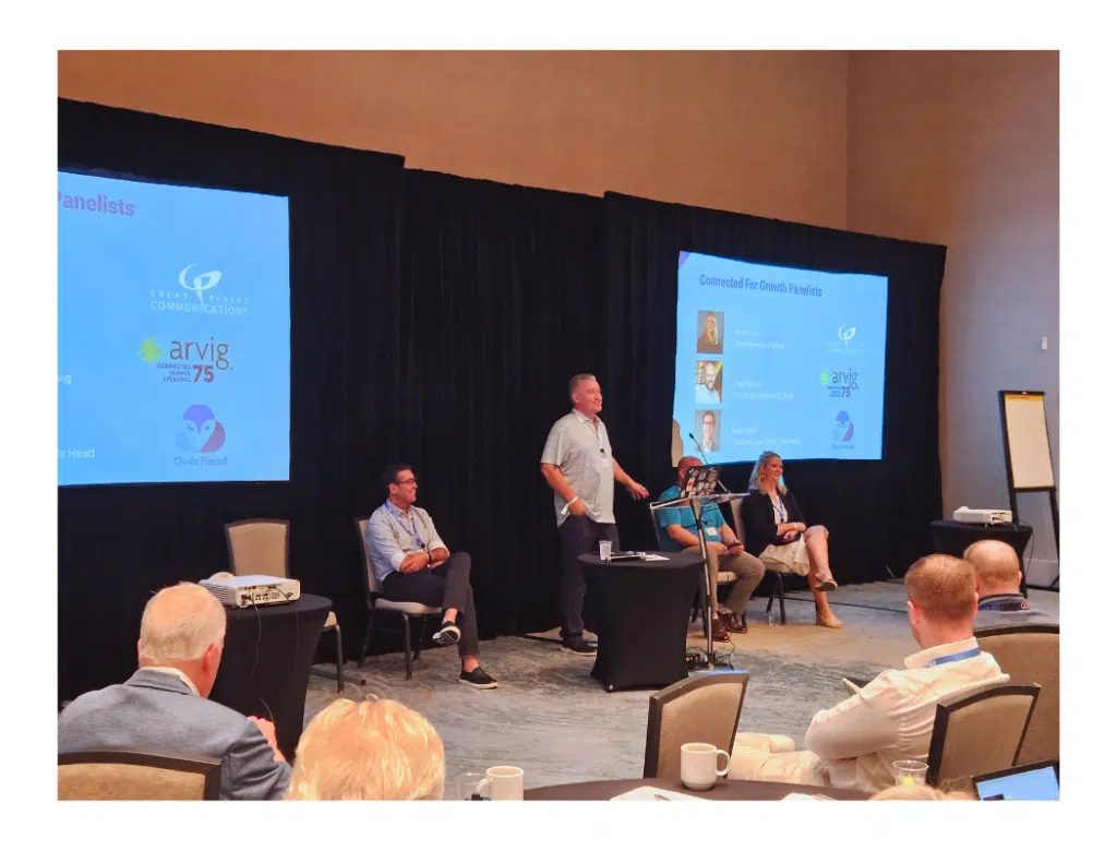 Man stands and speaks at a panel discussion on a stage, with three seated panelists and audience in the foreground; two blue slides read 'Connected For Growth Panelists'.