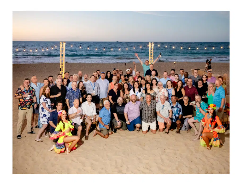 Large group of people on a sandy beach posing for a photo at dusk, with string lights and the ocean in the background.