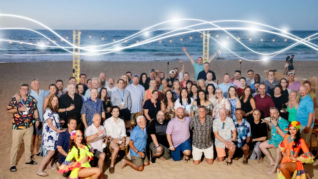 Large diverse group posing on a sandy beach with string lights and the ocean in the background, celebrating together.