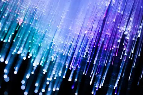 A close-up view of glowing fiber optic strands in shades of blue, purple, and teal, against a dark background, creating an abstract, futuristic effect.